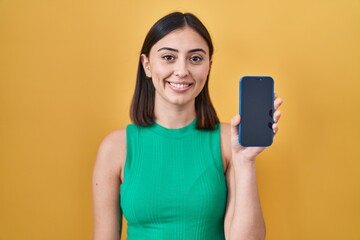 Naklejka premium Hispanic girl holding smartphone showing screen looking positive and happy standing and smiling with a confident smile showing teeth