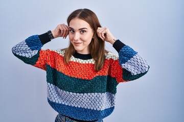 Young hispanic girl standing over blue background smiling pulling ears with fingers, funny gesture....