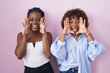 Two african women standing over pink background smiling cheerful playing peek a boo with hands showing face. surprised and exited