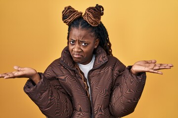 African woman with braided hair standing over yellow background clueless and confused expression...