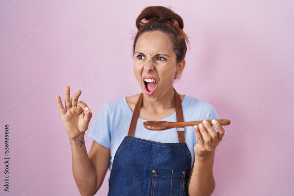 Poster Brunette woman wearing professional cook apron holding wooden spoon angry and mad screaming frustrated and furious, shouting with anger. rage and aggressive concept.