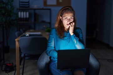 Brunette woman working at the office at night looking stressed and nervous with hands on mouth biting nails. anxiety problem.