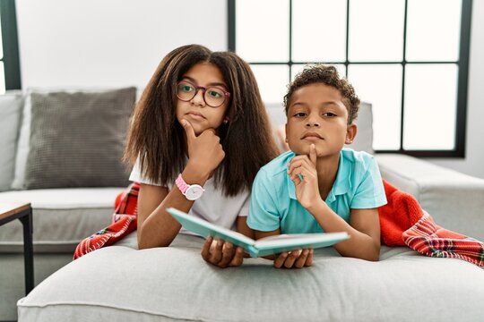 Two Siblings Lying On The Sofa Reading A Book Thinking Concentrated About Doubt With Finger On Chin And Looking Up Wondering