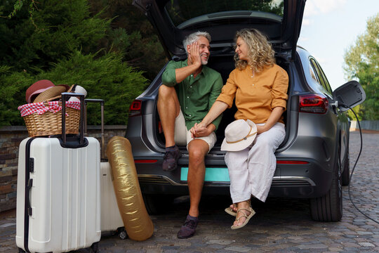 Middle-aged couple sitting in trunk while waiting for charging car before travelling on summer holiday.
