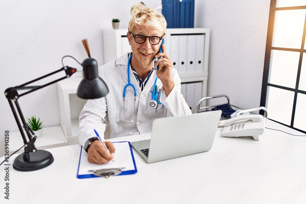 Poster Young caucasian man wearing doctor uniform talking on the smartphone at clinic