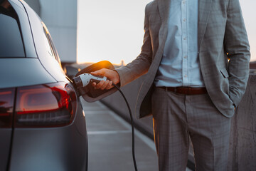 Businessman, holding power supply and charging his electric car during sunset. Concept of ecology...