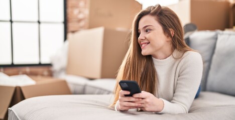 Young blonde woman using smartphone lying on sofa at new home