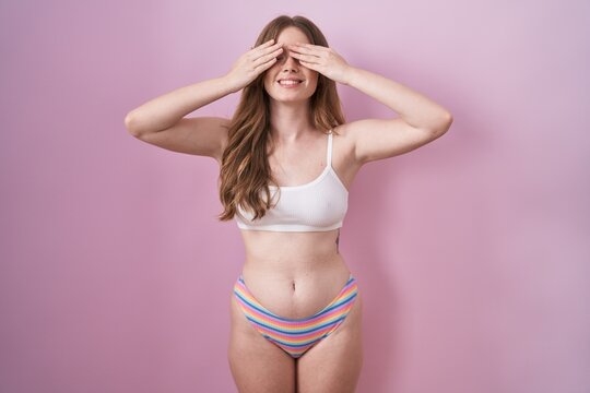 Caucasian woman wearing lingerie over pink background covering eyes with hands smiling cheerful and funny. blind concept.