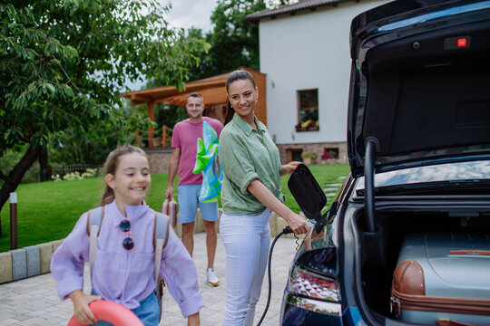 Happy mother holding power supply cable and charging their electric car, rest of family putting suitcases in car trunk, preparating for holidays.