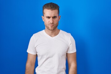 Young caucasian man standing over blue background skeptic and nervous, frowning upset because of...