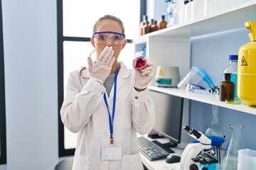 Young woman working at scientist laboratory holding geode covering mouth with hand, shocked and afraid for mistake. surprised expression