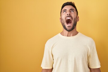 Handsome hispanic man standing over yellow background angry and mad screaming frustrated and...