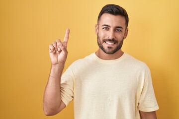 Fototapeta premium Handsome hispanic man standing over yellow background showing and pointing up with finger number one while smiling confident and happy.