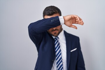 Handsome hispanic man wearing suit and tie covering eyes with arm, looking serious and sad. sightless, hiding and rejection concept