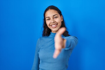 Fototapeta premium Young brazilian woman standing over blue isolated background smiling friendly offering handshake as greeting and welcoming. successful business.