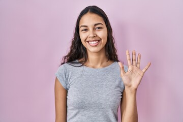 Young brazilian woman wearing casual t shirt over pink background showing and pointing up with fingers number five while smiling confident and happy.