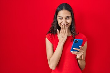 Young brazilian woman using smartphone over red background laughing and embarrassed giggle covering...
