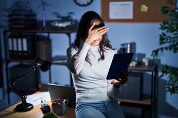 Young brazilian woman using touchpad at night working at the office covering eyes with hand,...