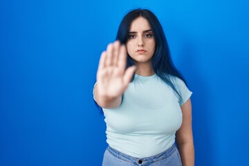 Young modern girl with blue hair standing over blue background doing stop sing with palm of the...
