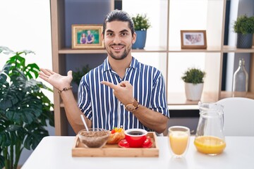 Obraz na płótnie Canvas Hispanic man with long hair sitting on the table having breakfast amazed and smiling to the camera while presenting with hand and pointing with finger.