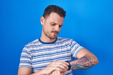 Young hispanic man standing over blue background checking the time on wrist watch, relaxed and confident