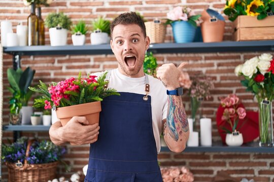 Young hispanic man working at florist shop holding plant pot pointing thumb up to the side smiling happy with open mouth