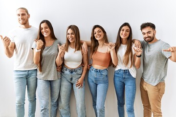 Group of young friends standing together over isolated background pointing to the back behind with hand and thumbs up, smiling confident