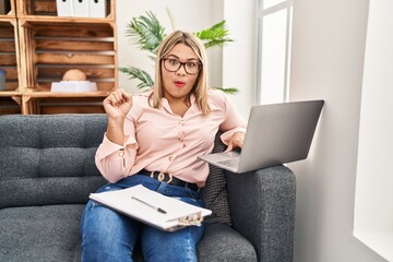 Young hispanic woman working online at consultation office scared and amazed with open mouth for surprise, disbelief face