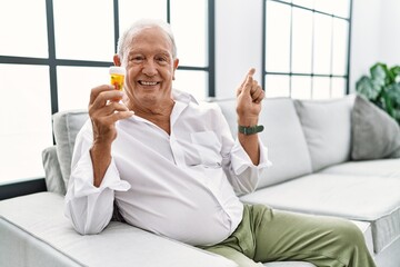 Senior man holding pills with a big smile on face, pointing with hand finger to the side looking at the camera.