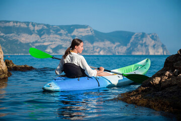 Girl swims in a kayak on the sea against the background of rocks near the shore