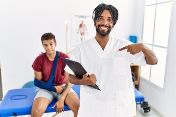 Young hispanic man working at pain recovery clinic with a man with broken arm looking confident with smile on face, pointing oneself with fingers proud and happy.