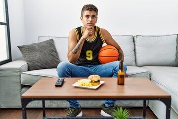 Young handsome hispanic man holding basketball ball cheering tv game serious face thinking about question with hand on chin, thoughtful about confusing idea