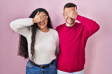 Obraz premium Young hispanic couple standing over pink background smiling and laughing with hand on face covering eyes for surprise. blind concept.