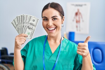 Young hispanic woman holding dollars banknotes working at pain recovery clinic smiling happy and positive, thumb up doing excellent and approval sign