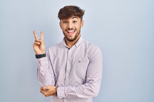 Arab man with beard standing over blue background smiling with happy face winking at the camera doing victory sign with fingers. number two.