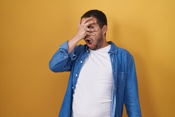 Hispanic man standing over yellow background peeking in shock covering face and eyes with hand, looking through fingers with embarrassed expression.