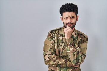Arab man wearing camouflage army uniform looking confident at the camera smiling with crossed arms and hand raised on chin. thinking positive.