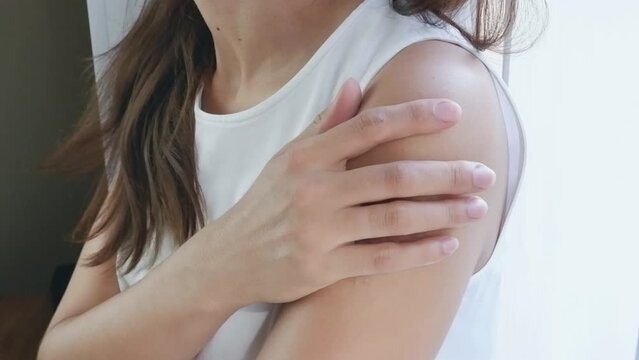 A woman in a white T-shirt by the window touches a large bruise on her arm with her hand. Domestic violence concept.