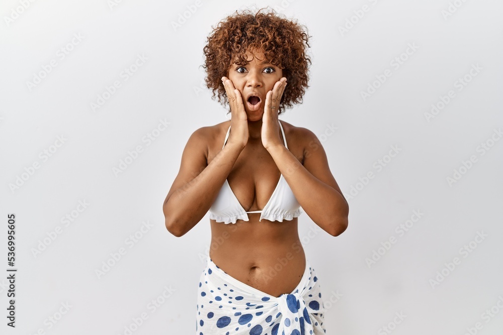 Canvas Prints Young african american woman with curly hair wearing bikini afraid and shocked, surprise and amazed expression with hands on face