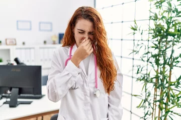 Foto op Canvas Young redhead woman wearing doctor uniform and stethoscope at the clinic smelling something stinky and disgusting, intolerable smell, holding breath with fingers on nose. bad smell © Krakenimages.com