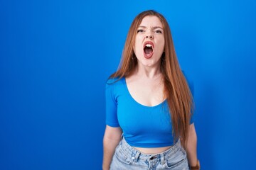 Redhead woman standing over blue background angry and mad screaming frustrated and furious, shouting with anger. rage and aggressive concept.