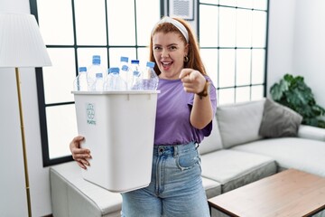 Fototapeta na wymiar Young redhead woman holding recycling wastebasket with plastic bottles pointing fingers to camera with happy and funny face. good energy and vibes.
