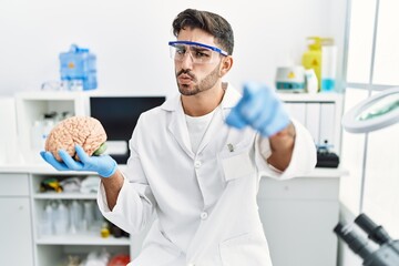 Young hispanic man working at laboratory holding medical brain pointing with finger to the camera and to you, confident gesture looking serious