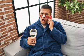 Hispanic young man wearing bathrobe and eye bags patches drinking wine speaking on the phone in...