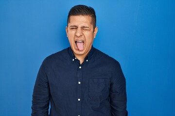 Hispanic young man standing over blue background sticking tongue out happy with funny expression. emotion concept.