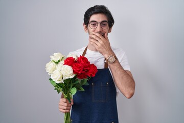 Young hispanic man holding bouquet of white and red roses laughing and embarrassed giggle covering mouth with hands, gossip and scandal concept