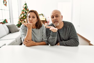 Middle age hispanic couple sitting on the table by christmas tree looking at the camera blowing a kiss with hand on air being lovely and sexy. love expression.