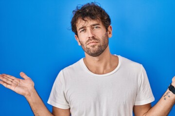 Hispanic young man standing over blue background clueless and confused expression with arms and...