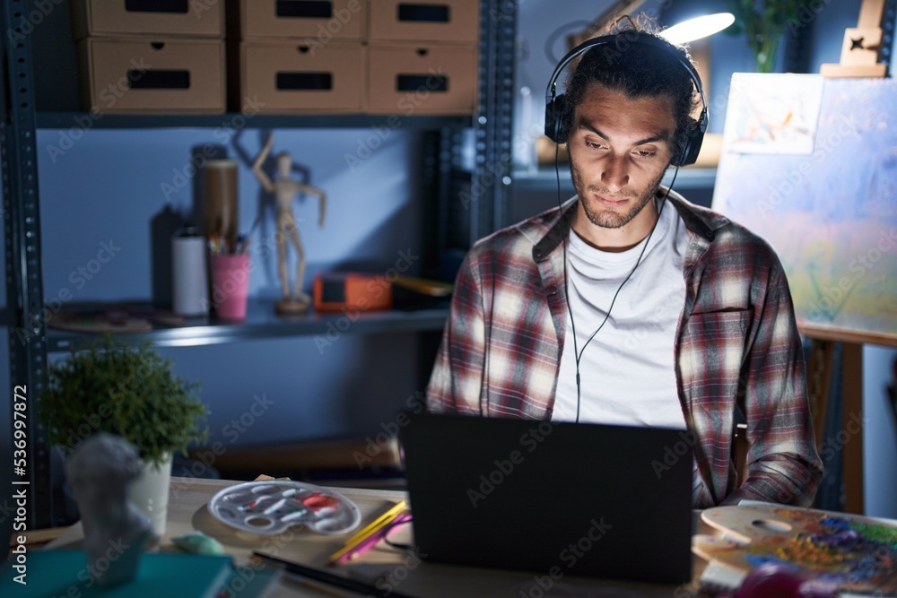 Poster Young hispanic man sitting at art studio with laptop late at night relaxed with serious expression on face. simple and natural looking at the camera.