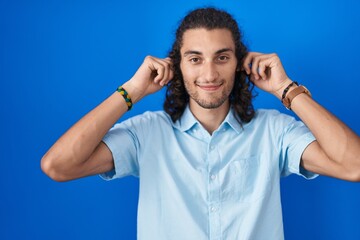 Young hispanic man standing over blue background smiling pulling ears with fingers, funny gesture....